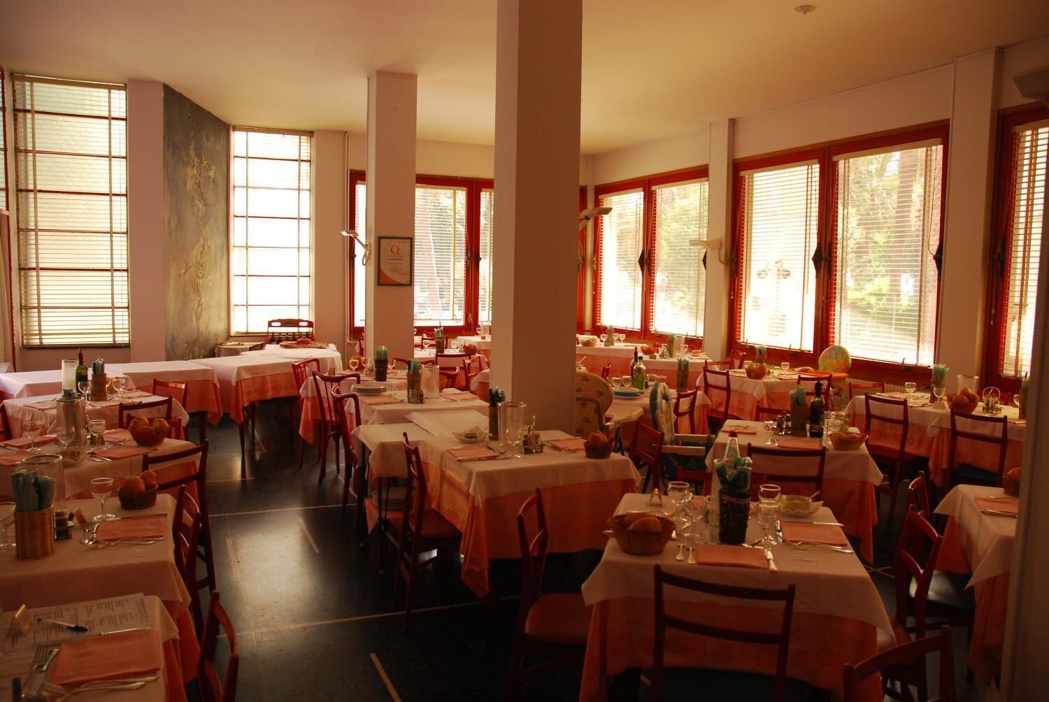 A restaurant with multiple tables set for dining, large windows with blinds, and a warm interior lighting.