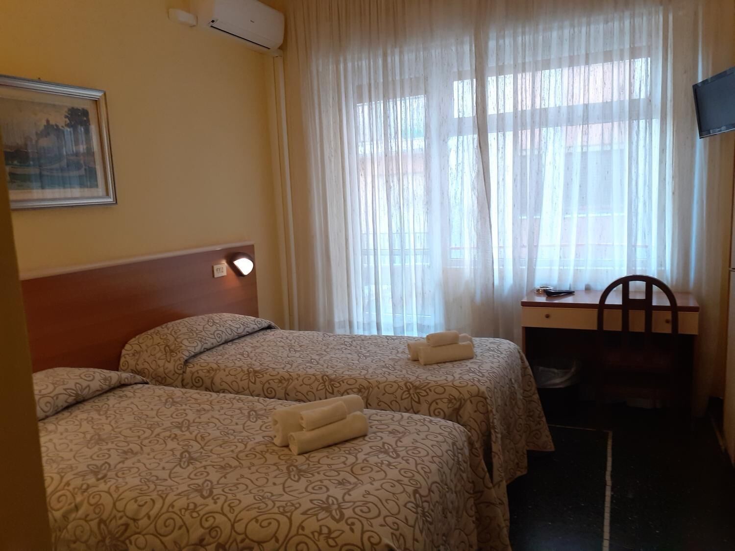 Et hotelværelse med to senge, et skrivebord, en stol, et fjernsyn og et vindue med gardiner.