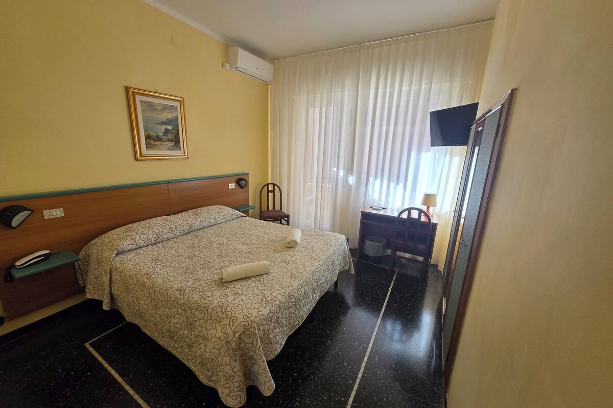 A hotel room with a bed, a desk, a chair, a TV, and a painting on the wall.