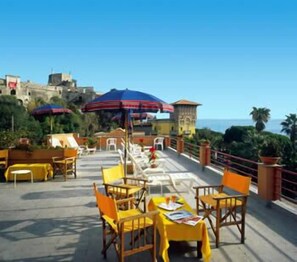 Zona pranzo all'aperto con tavoli e sedie gialle, un ombrellone blu e vista sul mare.