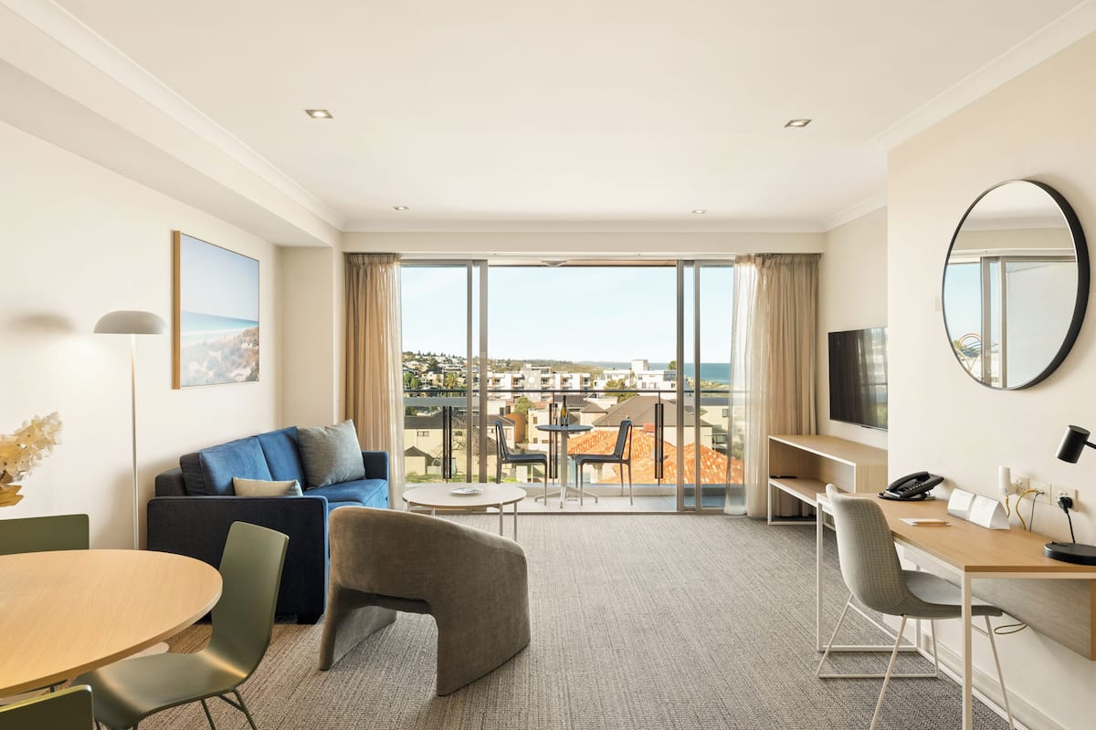 A modern hotel room with a large window, a sofa, a desk, and a dining table.