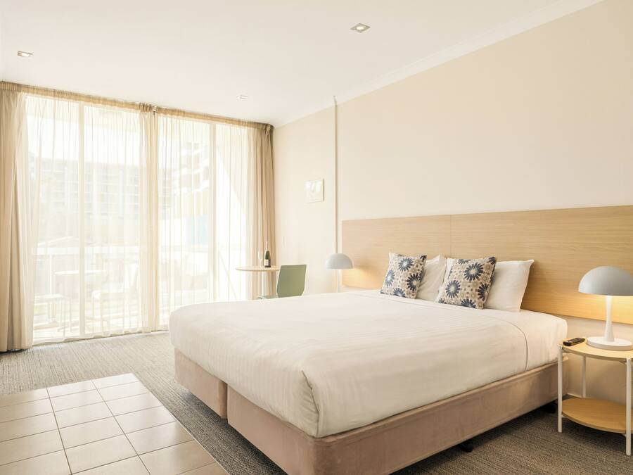 Habitación de hotel con una cama grande, dos mesitas de noche, una lámpara, una silla y vistas al exterior a través de cortinas translúcidas.