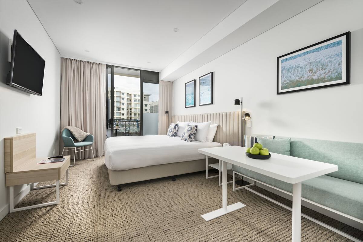 A modern hotel room with a bed, a desk, a sofa, a TV, and a window with a view of buildings.