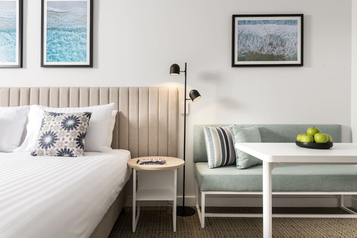 A hotel room with a bed, a bedside table with a magazine, a floor lamp, a sofa, and a small table with a bowl of apples.