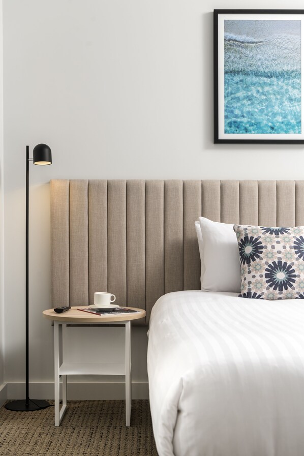 Een netjes ingerichte hotelkamer met een bed, nachtkastje met beker en afstandsbediening, vloerlamp en een ingelijst schilderij aan de muur.