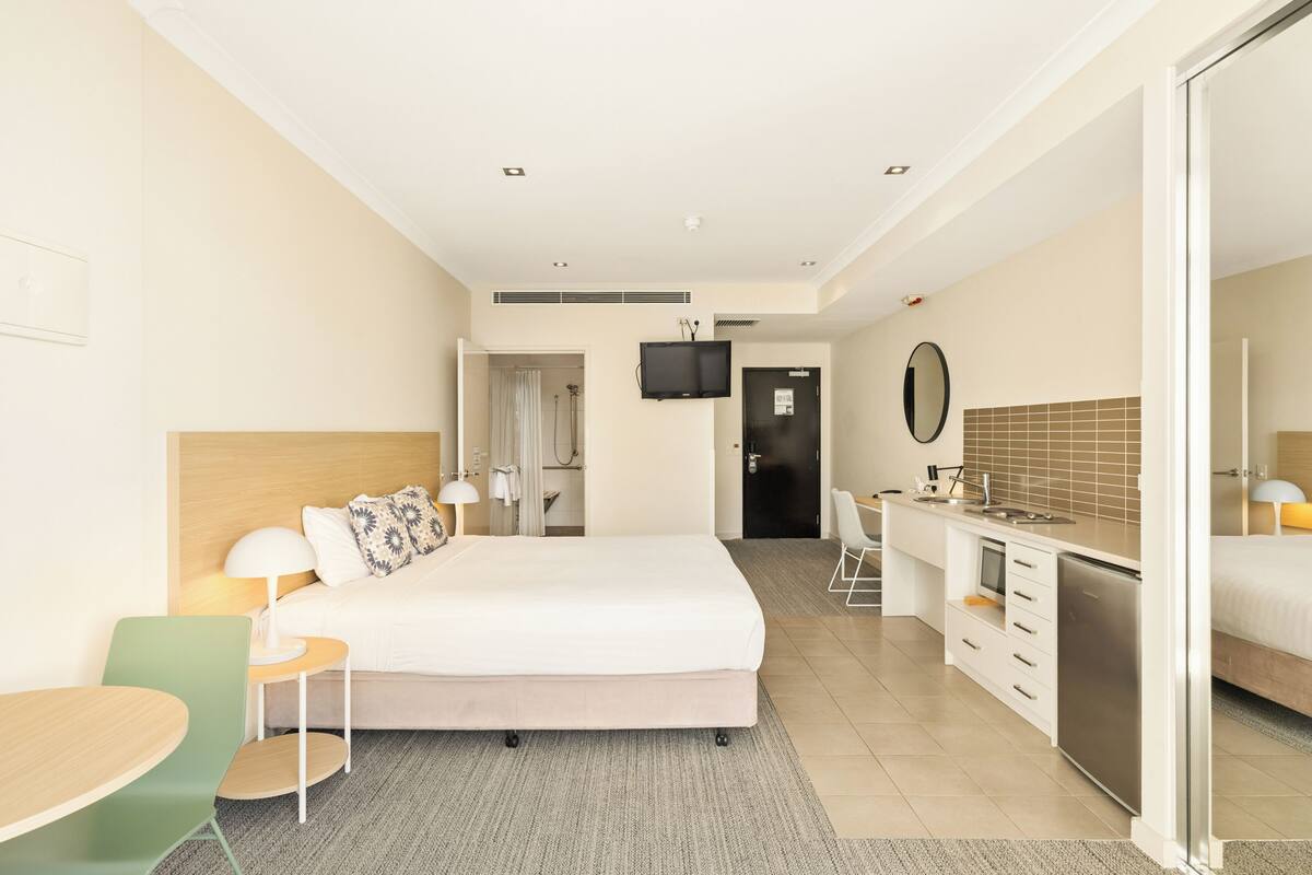 A hotel room with a bed, a desk, a chair, a TV, a mirror, and a kitchenette.