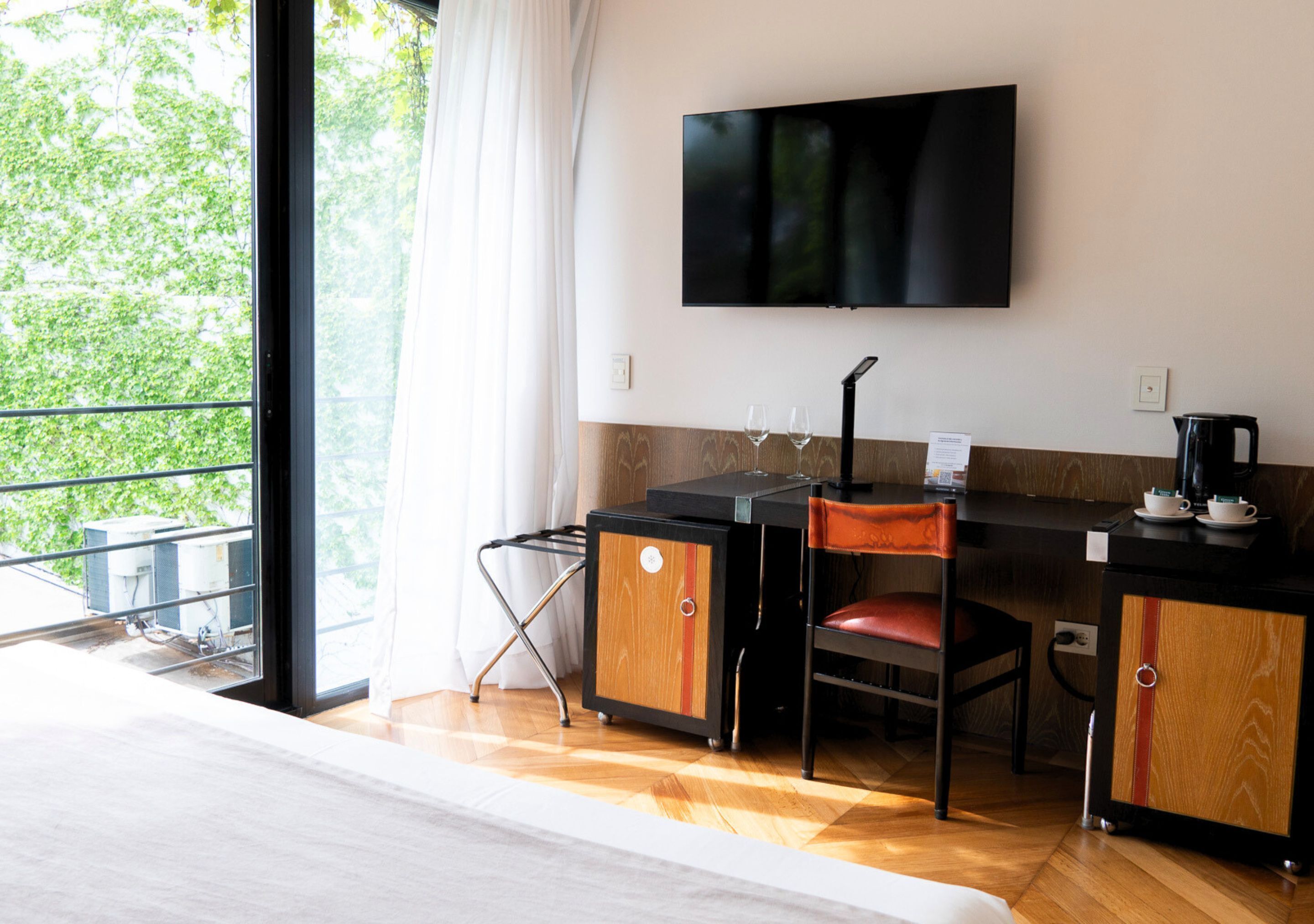 A hotel room with a bed, a desk with a chair, a television, and a view of greenery outside.