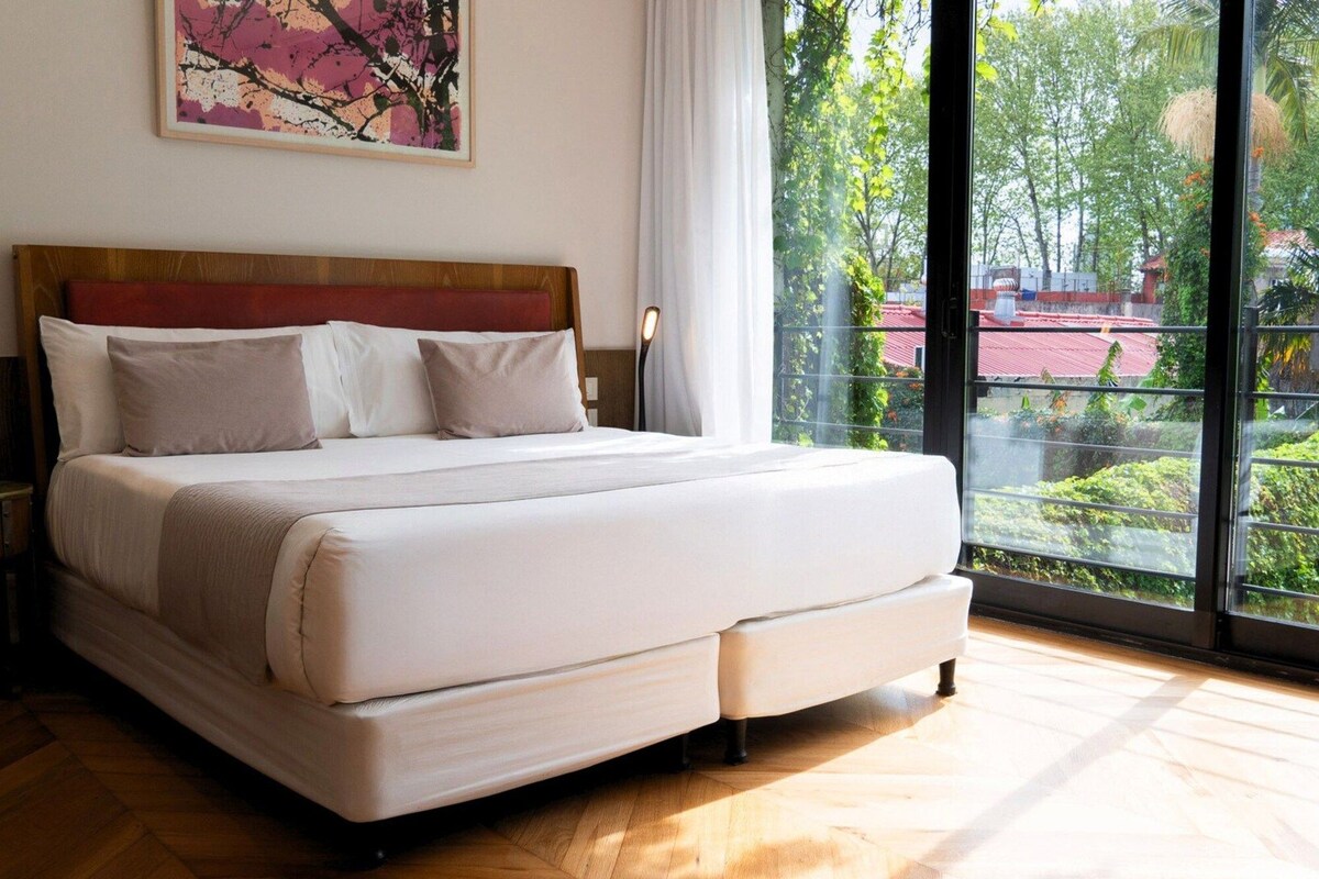 Una cama grande con cabecera de madera, ropa de cama blanca y vistas a la vegetación a través de un ventanal.