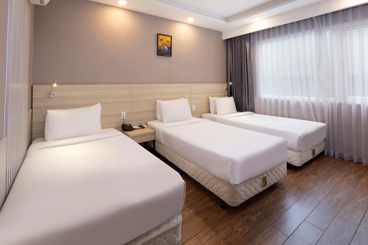 Une chambre d’hôtel avec deux lits simples, une table de chevet avec une lampe, un tableau au mur et une grande fenêtre avec des rideaux.