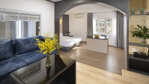 A modern hotel room with a sofa, a small table, a vase with flowers, a bed, a desk, and a view through an archway.