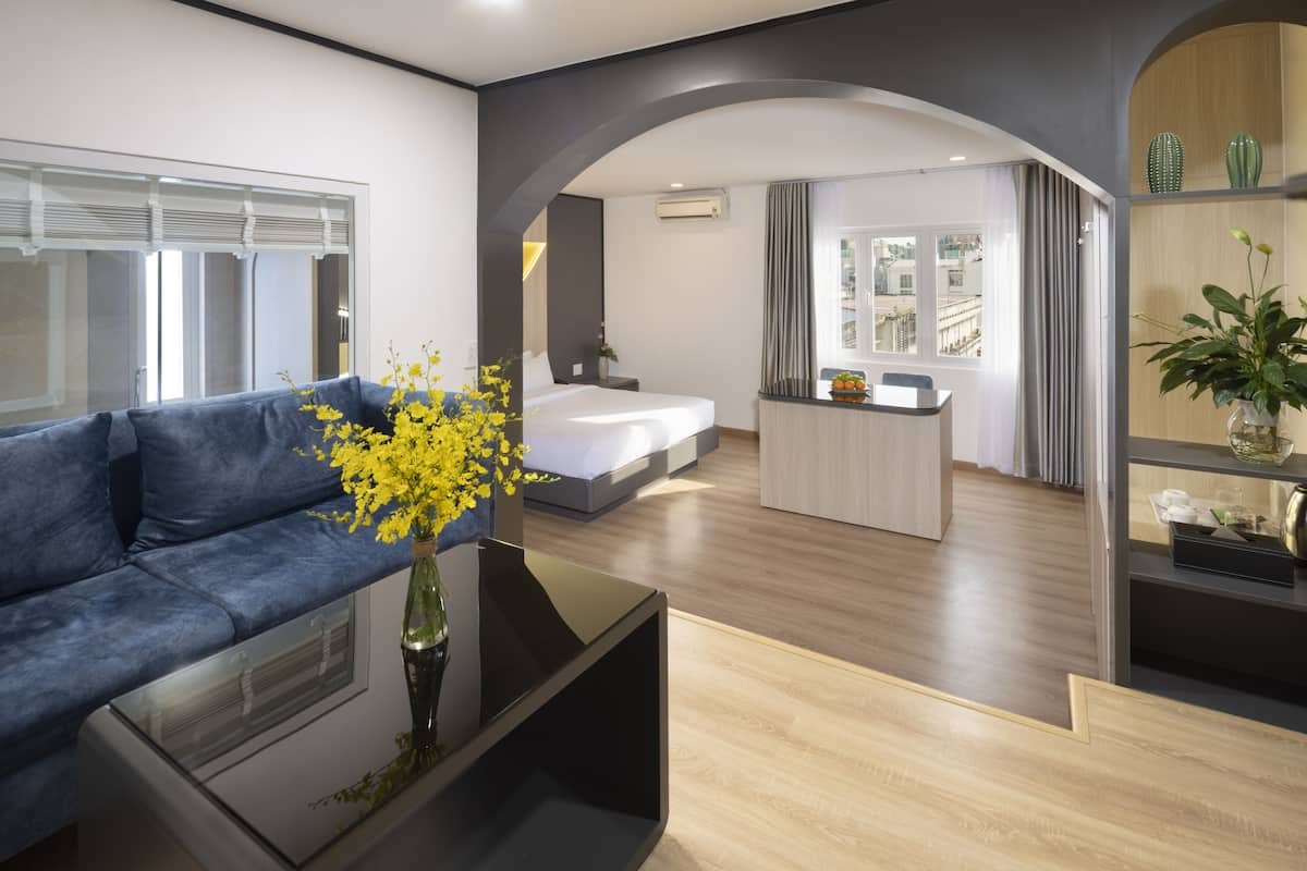 Une chambre d’hôtel moderne avec un canapé, une petite table, un vase de fleurs, un lit, un bureau et une vue sur l’extérieur, visible à travers une arche.
