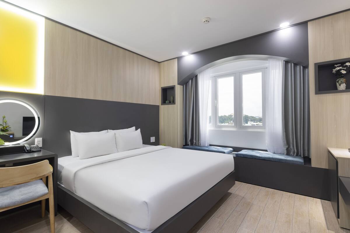 Une chambre d’hôtel moderne dotée d’un grand lit, d’une table de chevet avec une lampe, d’un bureau avec une chaise, d’une fenêtre avec des rideaux et d’une étagère murale agrémentée d’objets décoratifs.