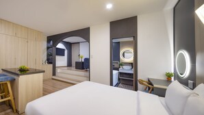 A modern hotel room with a bed, a desk with a chair, a small kitchenette, and a bathroom with a mirror and sink.