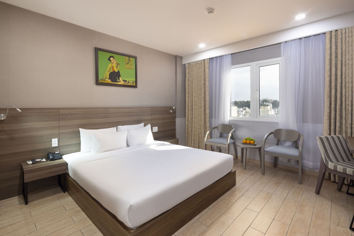 Une chambre d’hôtel avec un grand lit, un bureau avec un téléphone et une lampe, une chaise, une petite table avec des fruits et une fenêtre avec des rideaux.