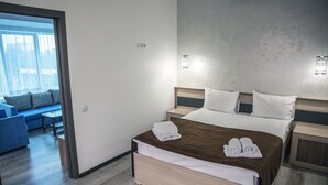 A modern hotel room with a large bed, a bedside table, a nightstand, a seating area with a sofa and a small table, and a view through a glass door. - Astra (Almaty)