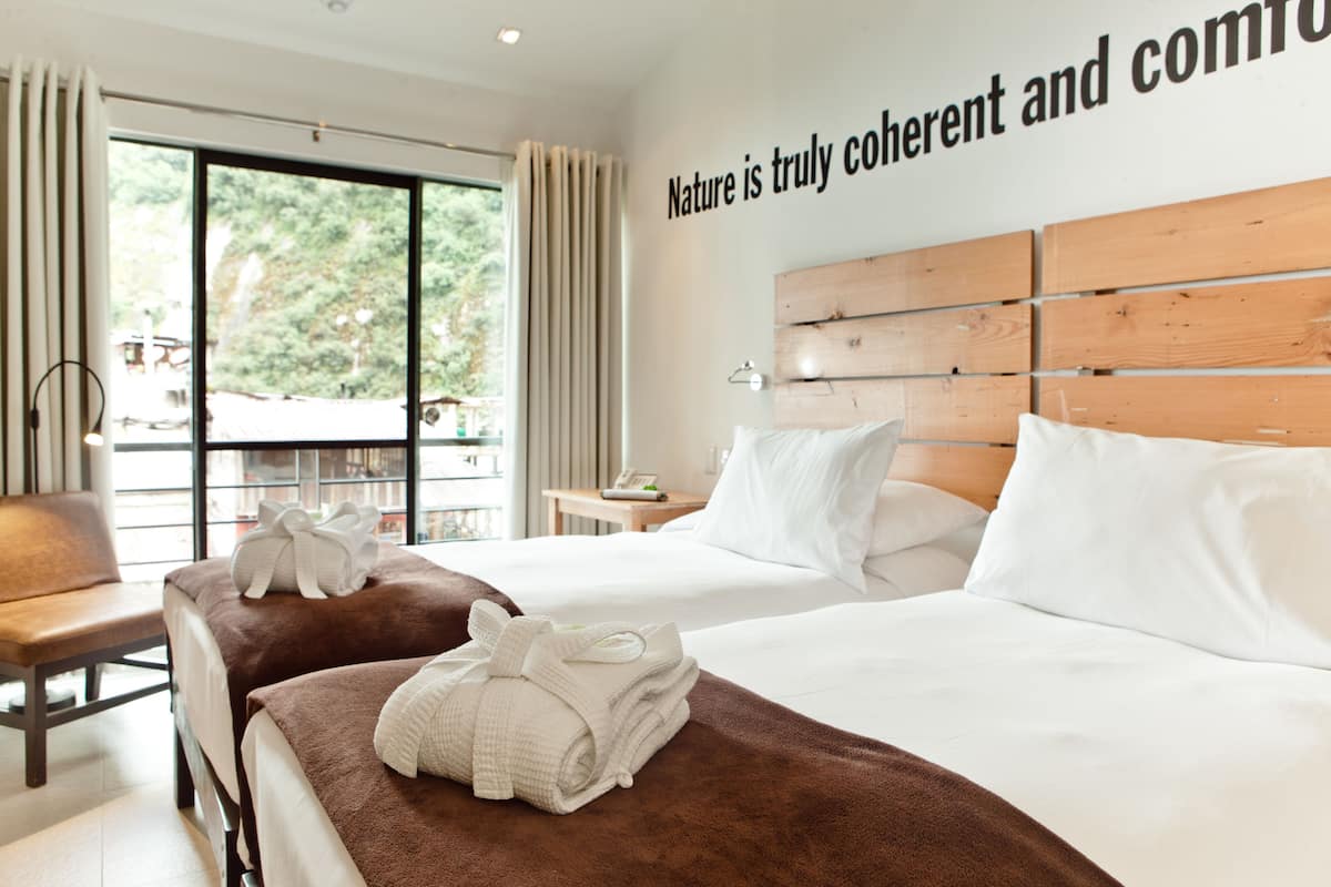 Habitación de hotel con dos camas, cabecero de madera, un ventanal grande con cortinas y una pared con una cita sobre la naturaleza.