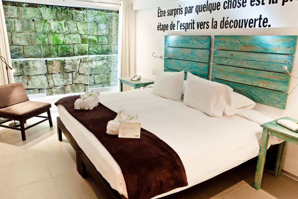 A hotel room with a large bed, a wooden headboard, a stone wall, a window, and a brown bench.