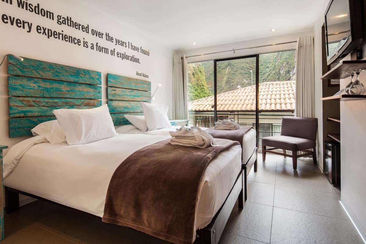 Habitación de hotel moderna con una cama grande, una cama más pequeña, una silla y un televisor. Se ve una ventana con vistas a un edificio y árboles.