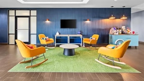 Un vestíbulo de hotel moderno con un televisor de pantalla plana, sillones roqueros anaranjados, una mesa redonda y un minibar azul.