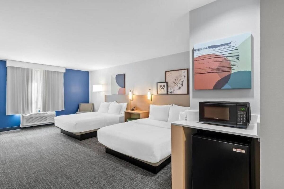 Habitación de hotel con dos camas, microondas, escritorio pequeño, mesita de noche y una obra de arte colgada en la pared.