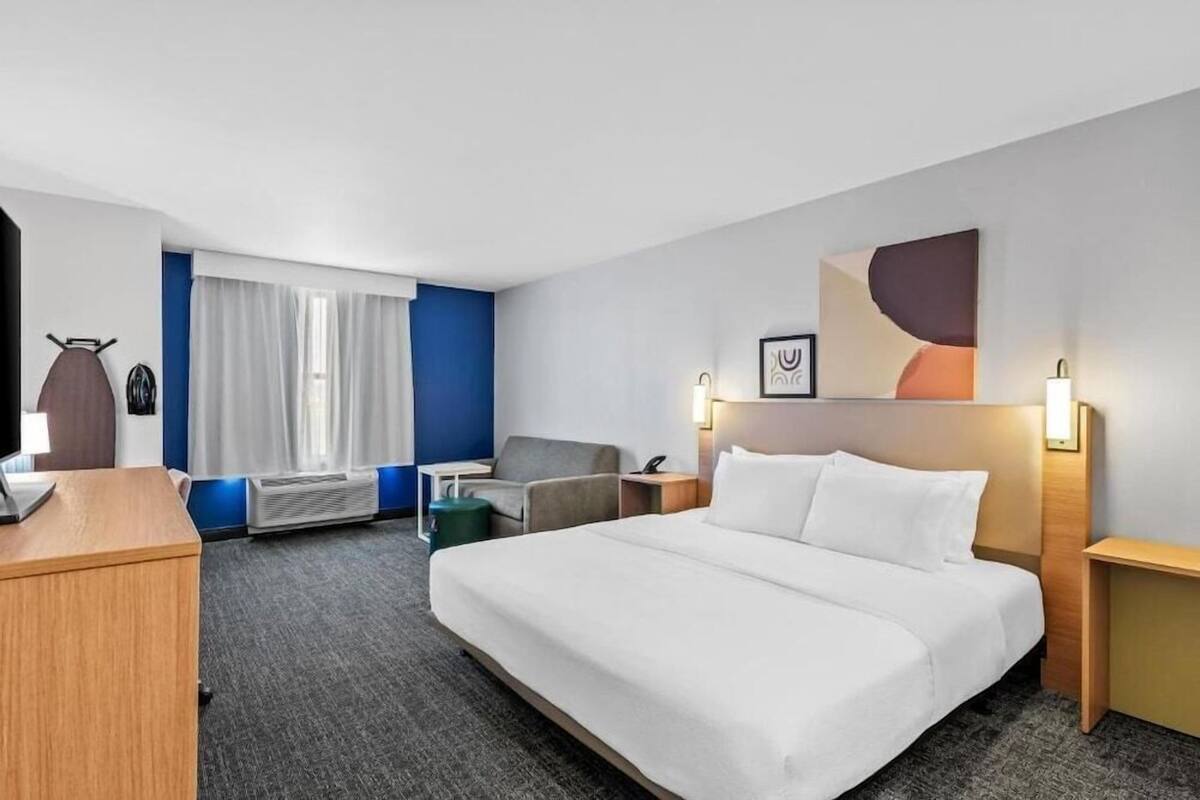 Habitación de hotel con cama, escritorio, silla, lámpara de escritorio, televisor montado en la pared y ventana con cortinas.