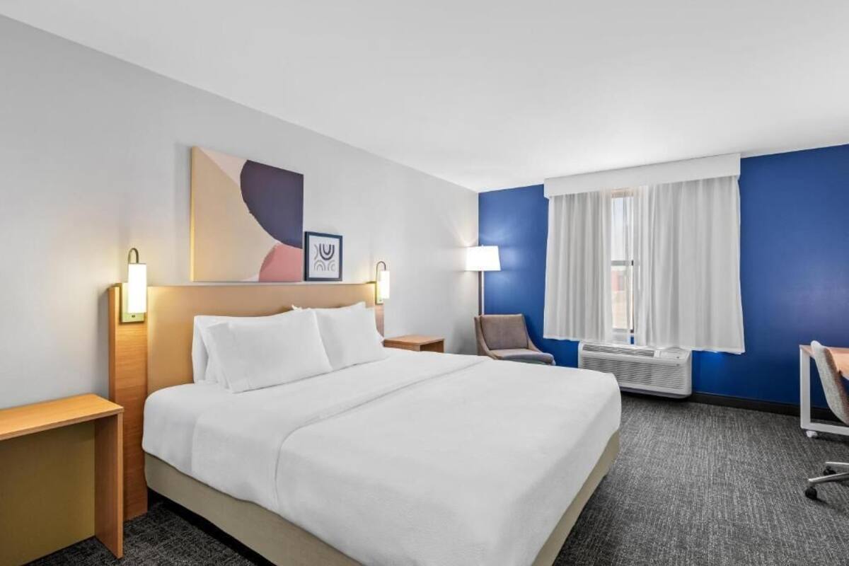 Habitación de hotel con una cama grande, mesitas de noche, una silla, un escritorio, un ventanal con cortinas y una obra de arte colgada en la pared.