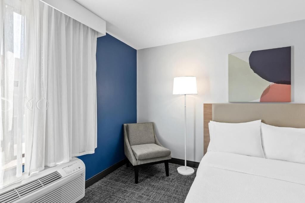 A hotel room with a bed, a chair, a floor lamp, and a window with curtains.