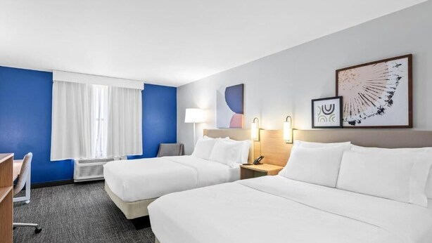 Habitación de hotel con dos camas, un escritorio, una silla, un ventanal con cortinas y decoración en la pared.