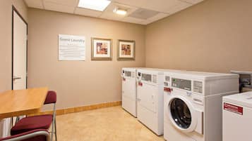 A laundry room with multiple washing machines, a table, chairs, and two framed pictures on the wall.