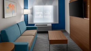 Una habitación de hotel moderna con una pared azul de acento, un sofá, una mesa de madera, un televisor de pantalla plana y una ventana con persianas.
