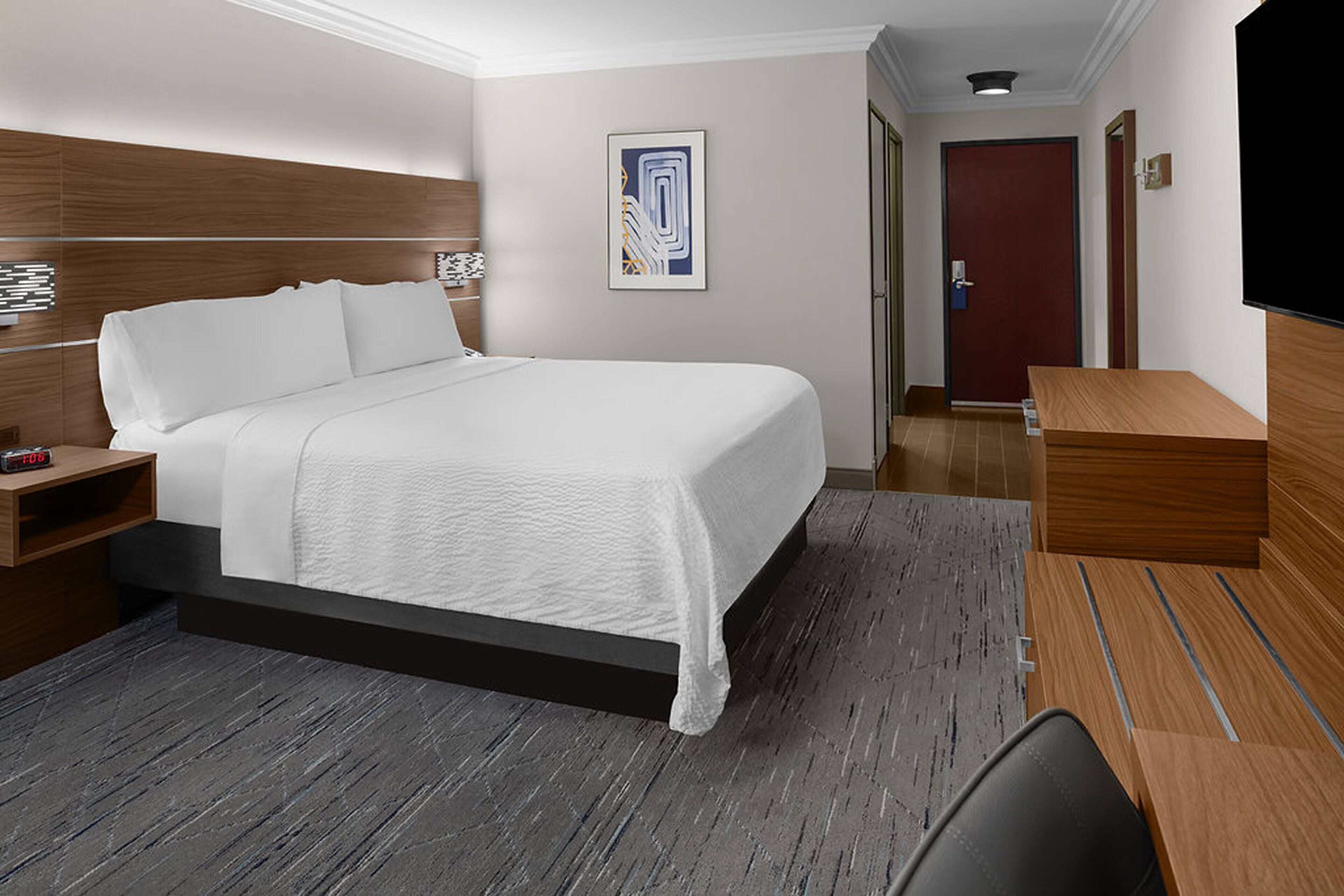 Quarto de hotel com uma cama grande, cabeceira de madeira, mesa de cabeceira com abajur, quadro na parede e TV fixada na parede.