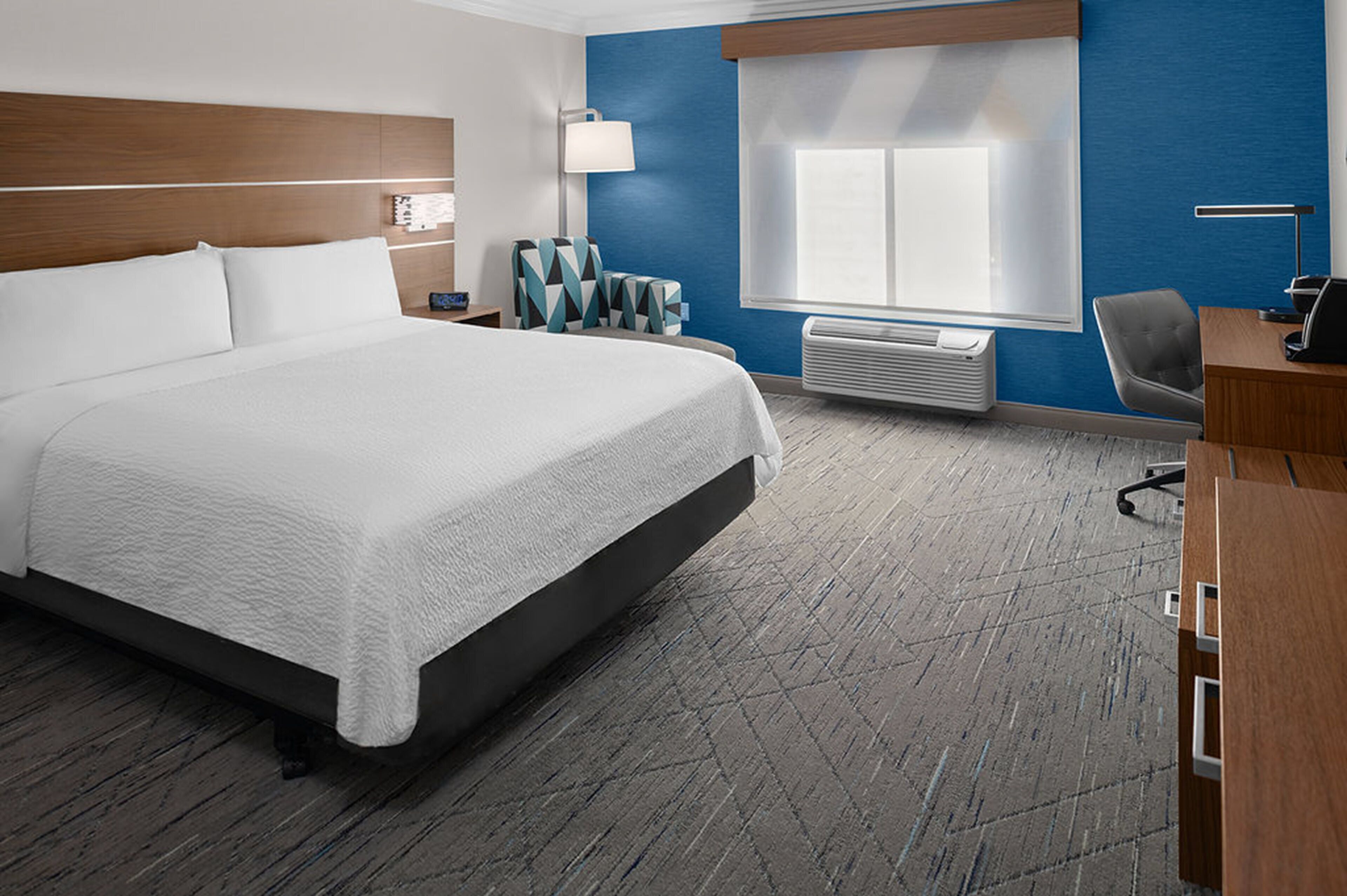 A hotel room with a large bed, a desk, a chair, a lamp, and a window with blinds.