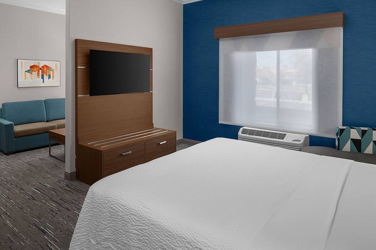 Une chambre d’hôtel avec un grand lit, une tête de lit en bois, un téléviseur à écran plat, une fenêtre avec des stores, un mur d’accent bleu, un canapé bleu et une petite table.