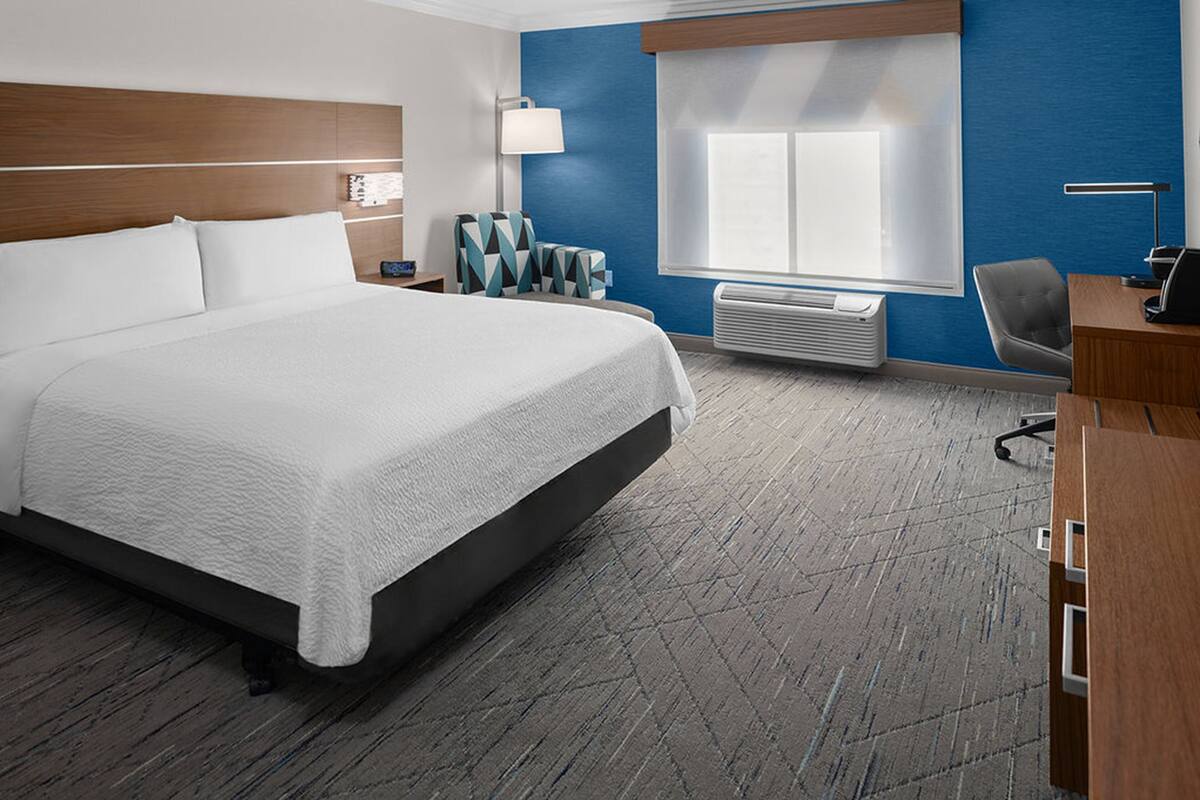 Une chambre d’hôtel avec un grand lit, un bureau, une chaise, une lampe et une fenêtre avec des stores.