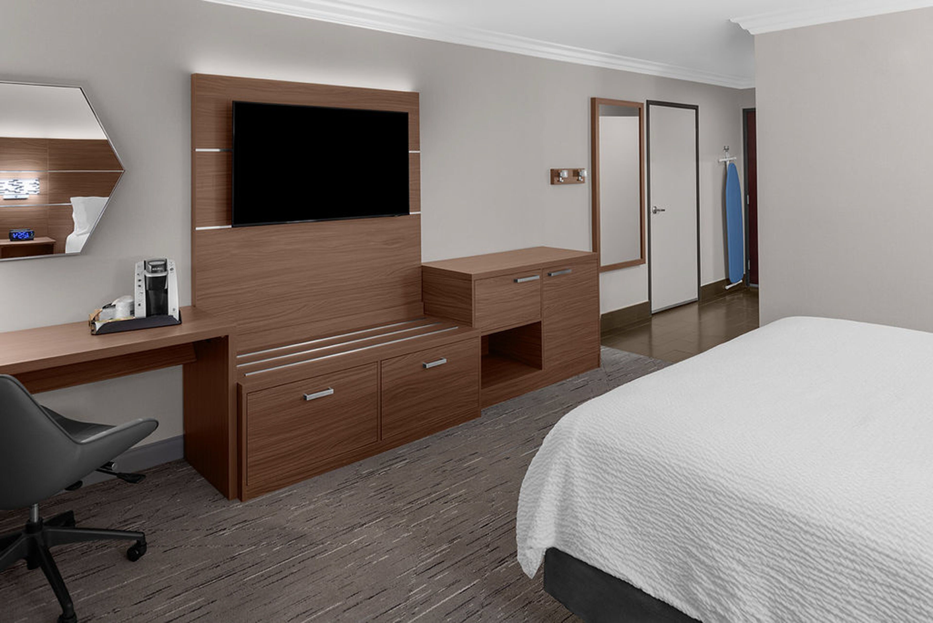 Habitación de hotel con un escritorio de madera, un televisor de pantalla plana, una cama con ropa de cama blanca, una silla y un espejo.