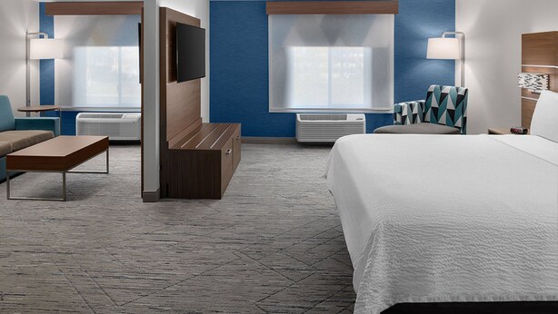 Habitación de hotel con una cama grande, dos sillones, un sofá, un televisor y un ventanal con persianas.