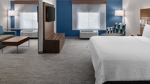 Habitación de hotel con una cama grande, dos sillones, un sofá, un televisor y un ventanal con persianas.