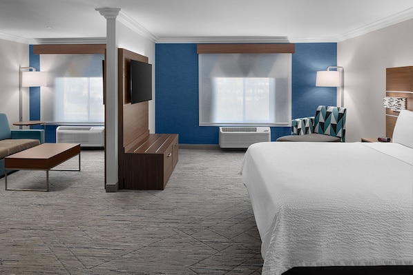 Habitación de hotel con una cama grande, dos sillones, un sofá, un televisor y un ventanal con persianas.