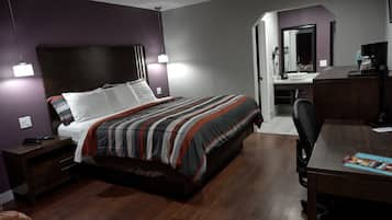 Habitación de hotel con cama, escritorio, silla, espejo y baño visible a través de una puerta abierta.