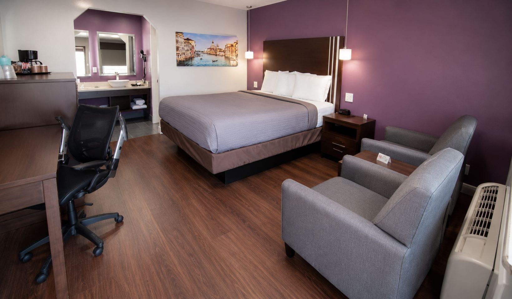 Ett hotellrum med en säng, ett skrivbord, en stol, en spegel och en tavla på väggen.