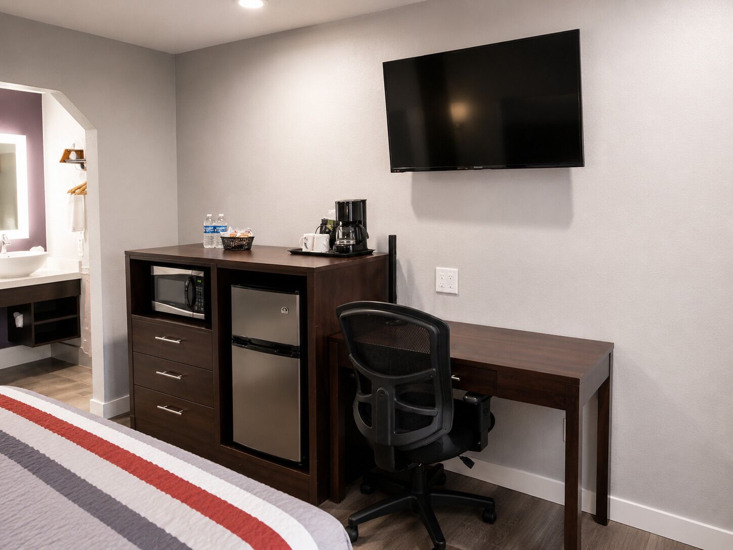 Quarto de hotel com cama, escrivaninha com cadeira, televisão, minibar e banheiro com pia e espelho.