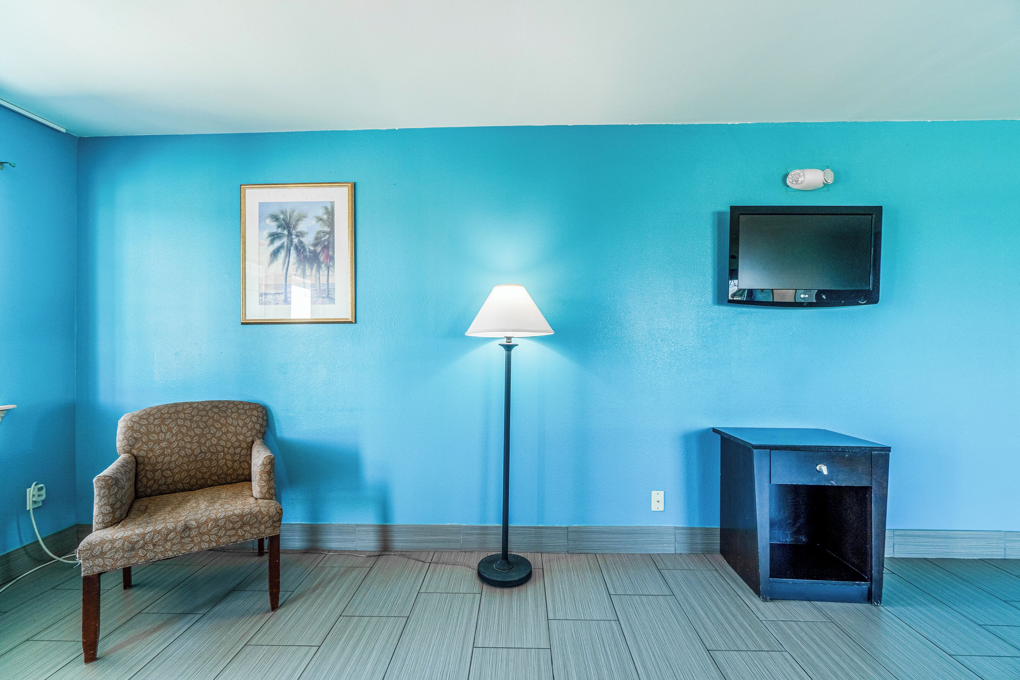 Una habitación con pared azul, un cuadro enmarcado, una lámpara de pie, un televisor, una silla y un armario pequeño.