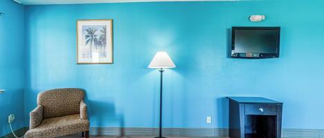 Una habitación con pared azul, un cuadro enmarcado, una lámpara de pie, un televisor, una silla y un armario pequeño.