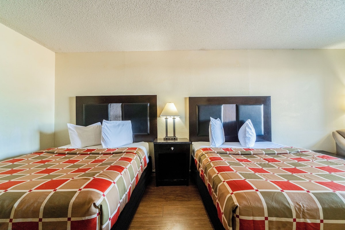 Quarto de hotel com duas camas, cada uma com cabeceira, mesa de cabeceira com abajur e piso de madeira.