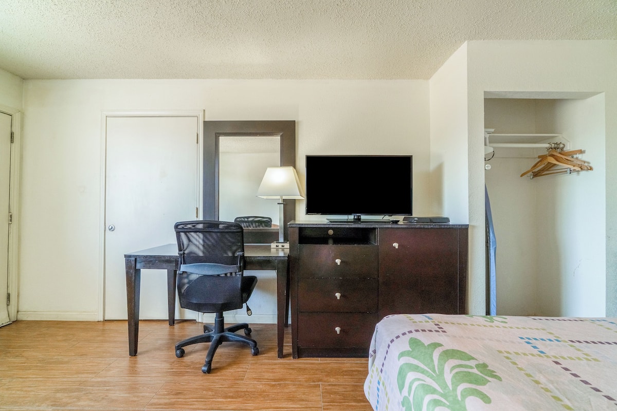 Quarto de hotel com escrivaninha de madeira, cadeira de escritório preta, televisão em uma cômoda, cama com colcha estampada e cabideiro com ganchos.