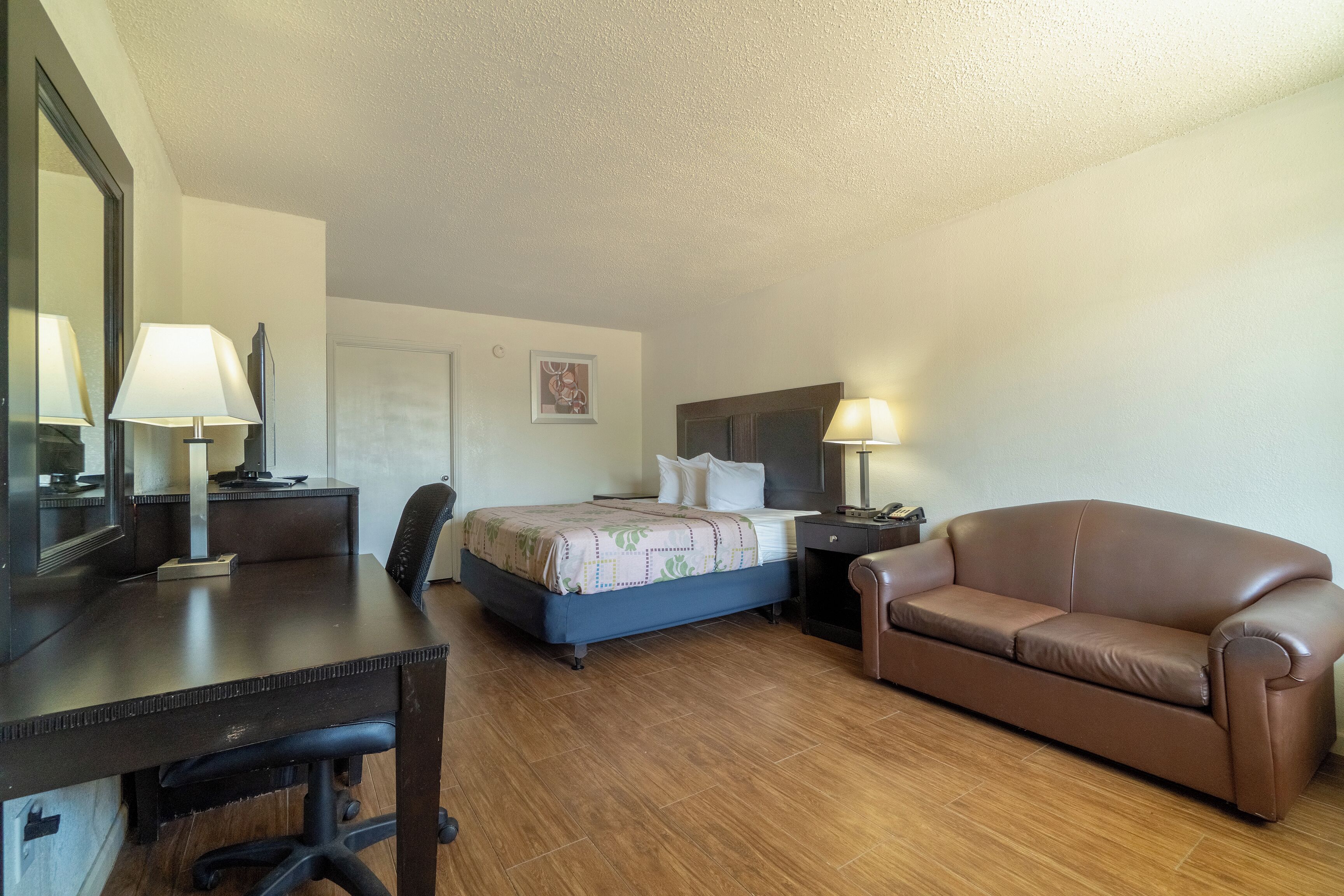 A hotel room with a bed, a desk, a chair, a sofa, and a nightstand with a lamp.