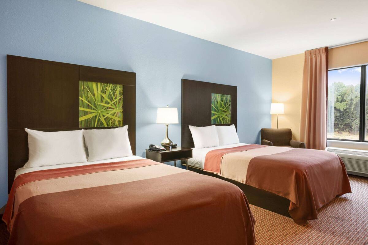 A hotel room with two beds, a nightstand, a lamp, and a chair.