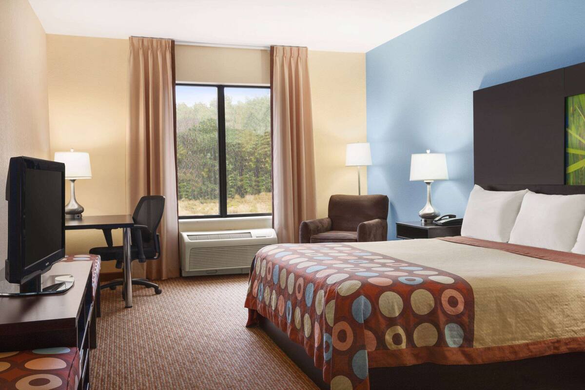A hotel room with a large bed, a desk with a computer, two chairs, a TV, and a window with curtains.