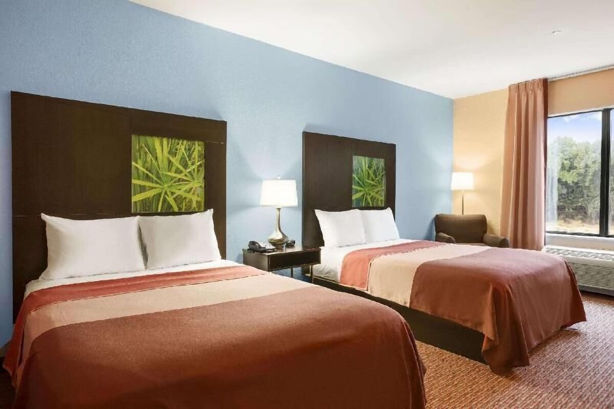 A hotel room with two beds, a nightstand with a lamp, a chair, and a large window with a view.