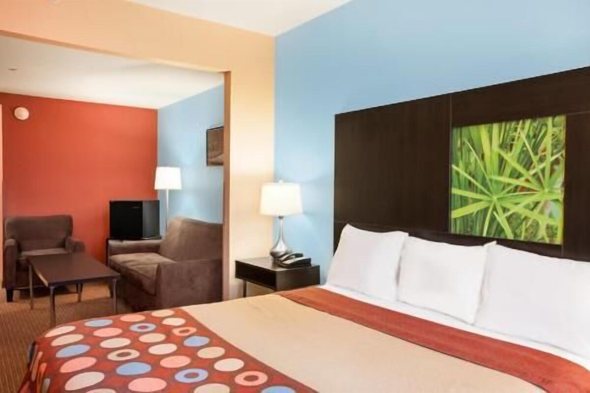 A hotel room with a bed, a desk, two chairs, a TV, and a nightstand with a lamp.